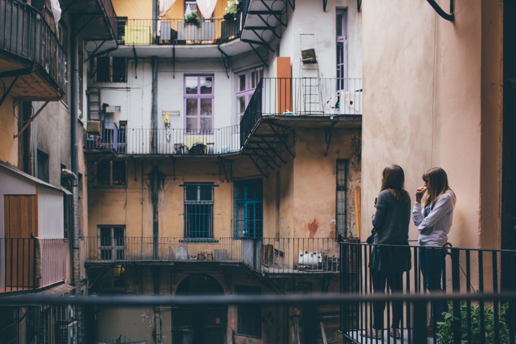 Two young women stand in patio together and look at it. Colorful windows in old city or town. Travel together